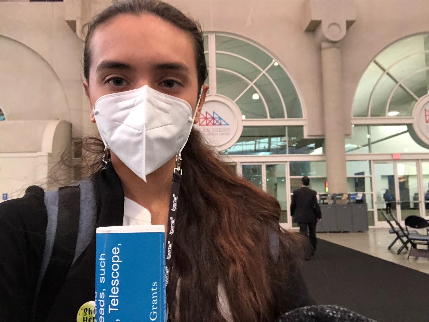 a woman with brown skin and dark hair is wearing professional clothing and a mask while holding a poster tube inside a conference room